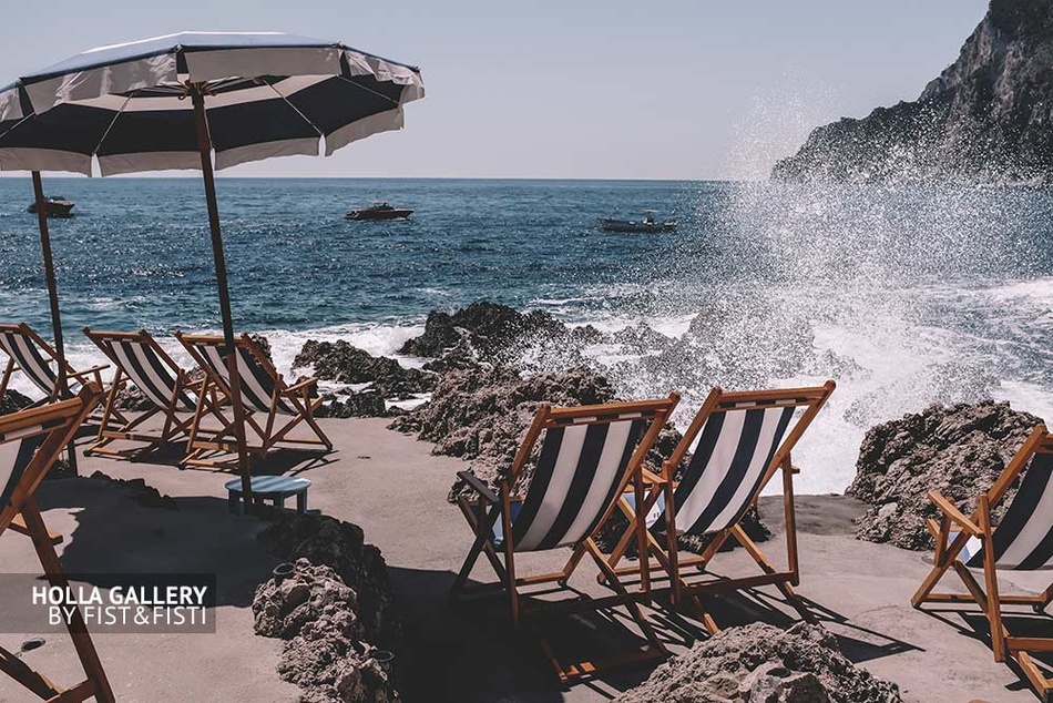 Брызги волн у каменистого пляжа с шезлонгами на Capri. На фоне лодки в заливе. Фото для интерьера